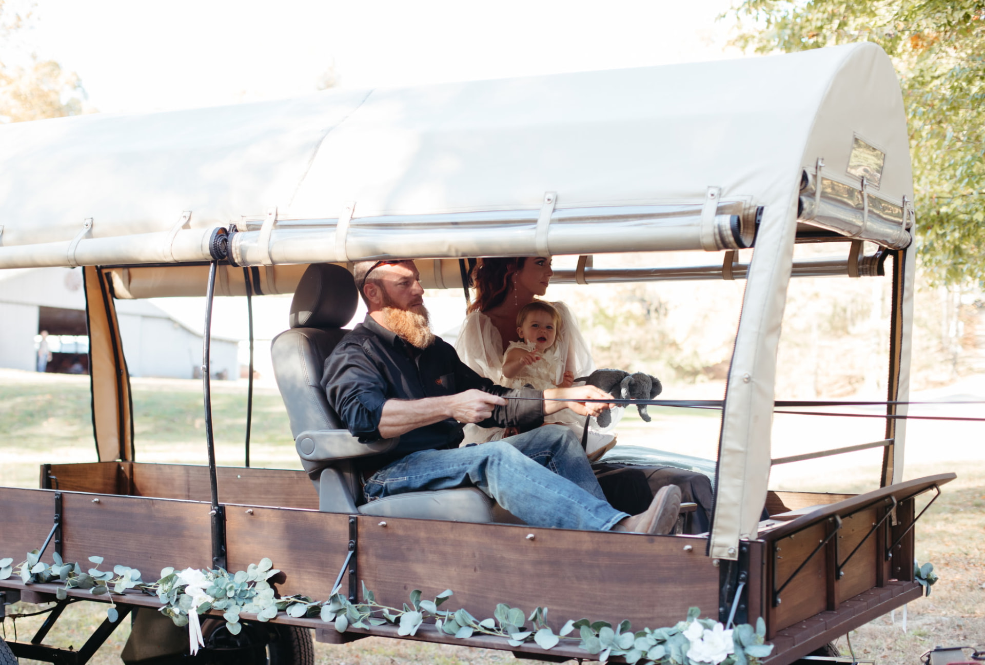 A bride and groom with their little one riding in the flower-trimmed canopy wagon on their wedding day